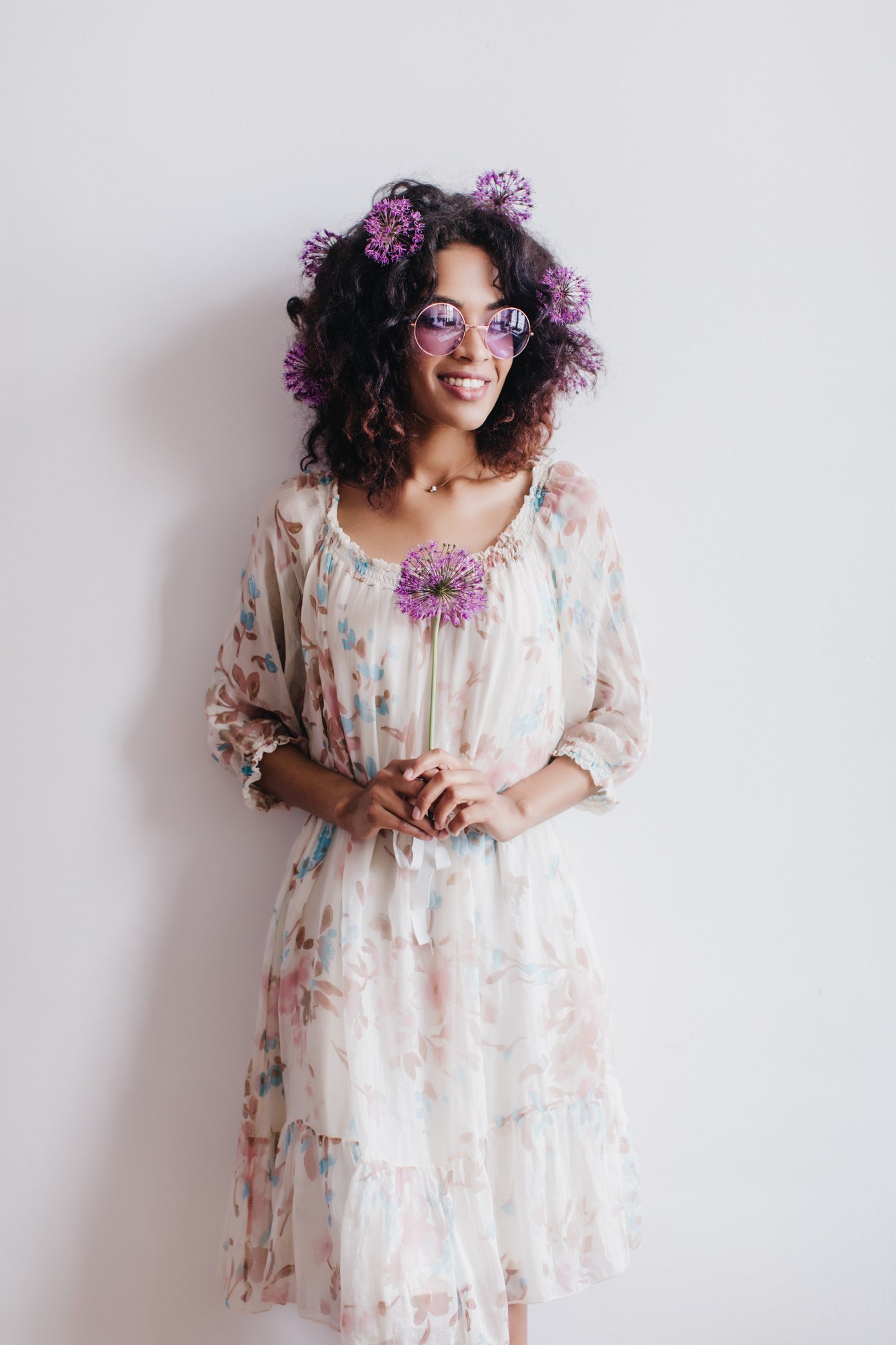 Wonderful Black Female Model Standing in Front of White Wall with Flower. Happy African Lady Holding Purple Allium during Indoor Photoshoot.